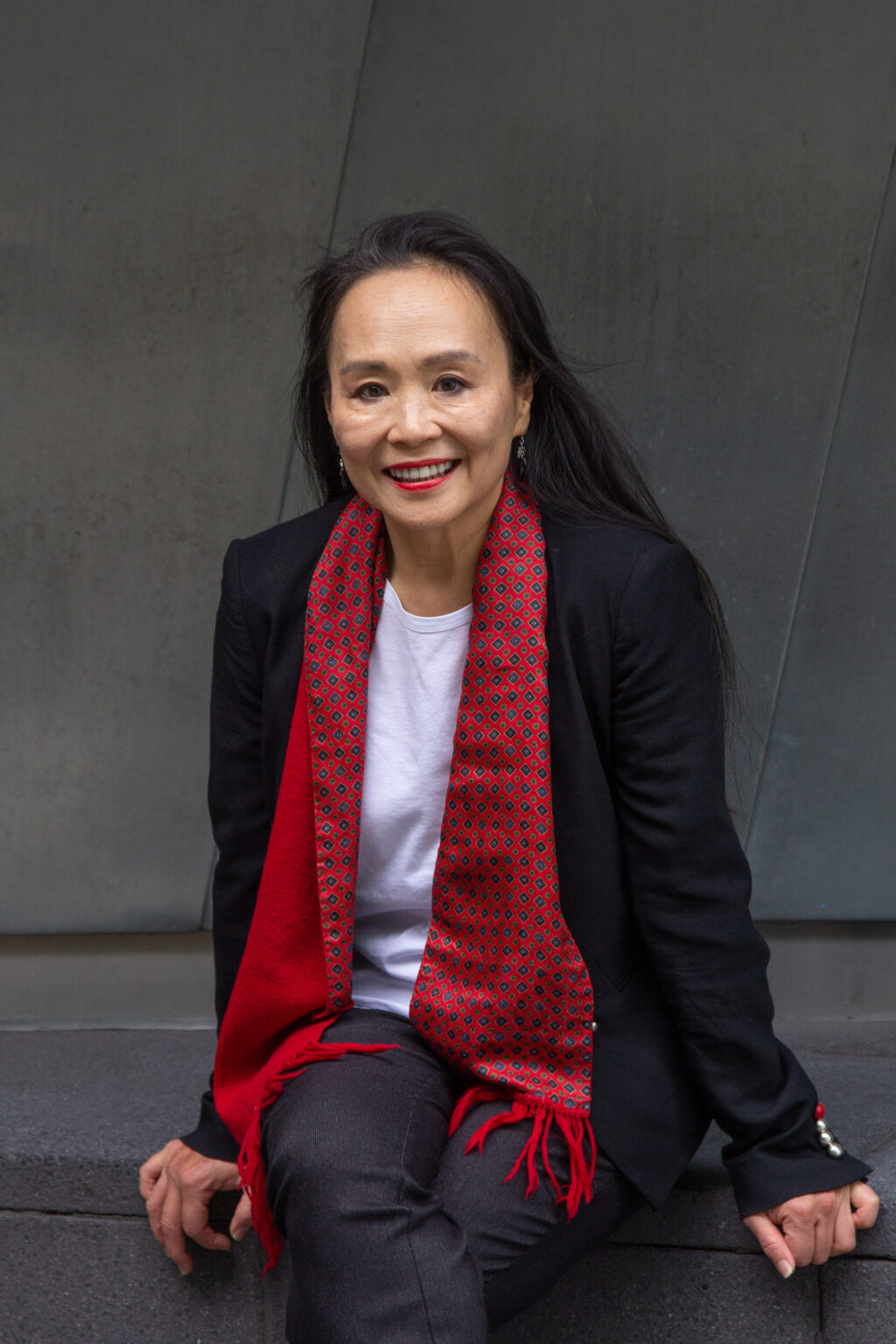Photography of Anne Pang wearing a black suit and red scarf, sitting.